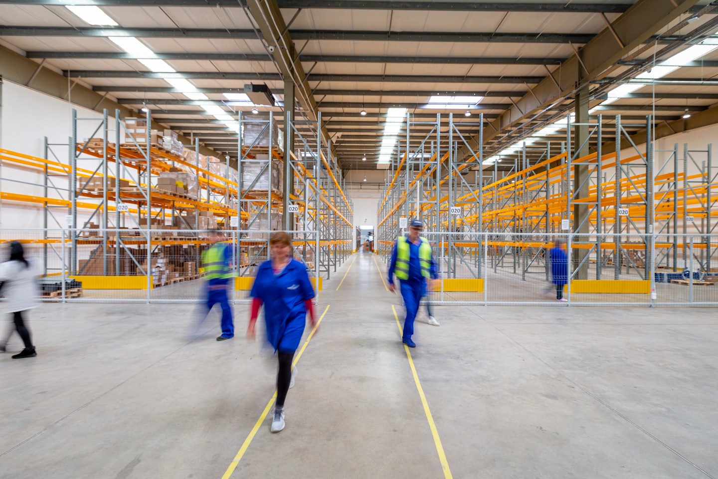 Magazijnmedewerkers in uniformen en hesjes lopen door een gang met opslagrekken.